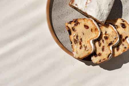 Stollen, fruit cake or bread with raisins and sugar powder sliced on pieces on a plate on neutral beige background with sunlight shadow, flat lay, copy space. Traditional German winter holiday bakery.の写真素材