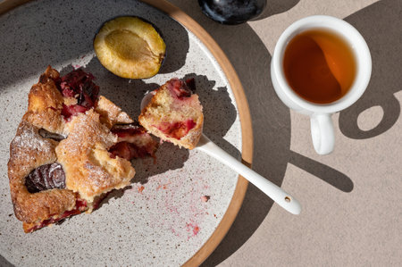 Piece of plum torte on a plate, cup with tea, teaspoon on neutral beige table with sunlight shadow. Lifestyle morning breakfast, sliced fruit pie, cakeの写真素材