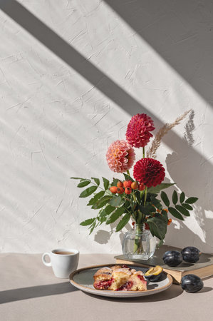 Sweet cozy autumn morning breakfast. Plum pie piece on plate, cup of tea, vase with autumn flower bouquet on table, aesthetic sunlight shadows on white wall backgroundの写真素材