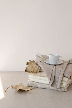 White coffee cup standing on stack of knitted beige pastel color sweaters, with dried fall leaves, on table. Empty neutral wall background, copy space.の写真素材