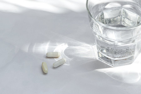 White pills and capsules, glass with pure water on white table background with natural sunlight shadows. Minimal aesthetic health care and medicine concept, close up.の写真素材