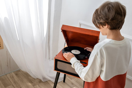 Beautiful teenager boy putting vinyl record in retro wooden brown turntable, player, preparing listening music. Back view, minimalist home interiorの写真素材