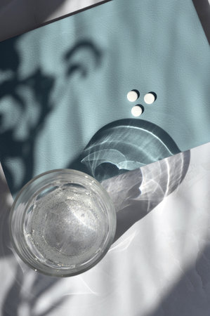 Aesthetic flat lay with pills in blue notebook, glass with water on white table background with...