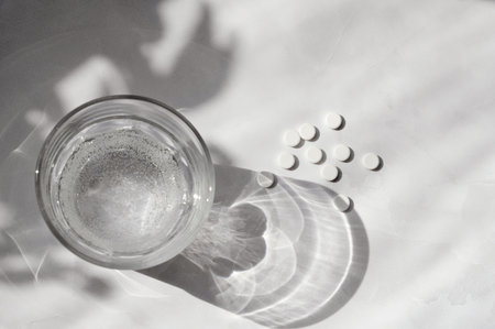White pills, glass with pure water on neutral white background with aesthetic sunlight shadow....