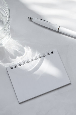 Empty paper notebook sheet mock up, pen, glass with water on white table background. Soft aesthetic natural sunlight shadows. Minimal business branding template.の写真素材