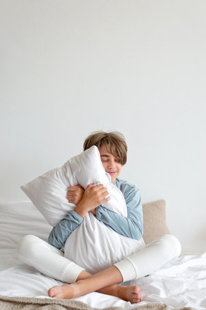 Good sleeping, time to sleep, youth daily health habit concept. Teenager boy sitting on bed, hugging pillow and smiling. Empty white wall background.の写真素材