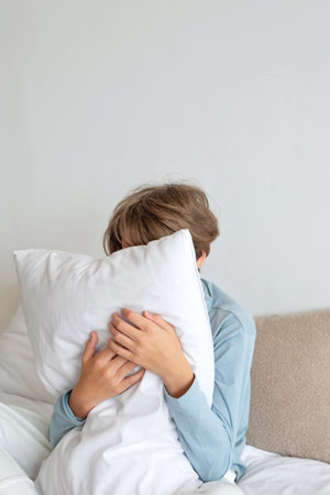 Teenager sleep deficiency, time to sleep, insomnia concept. Boy sitting on bed, embraicing pillow, hiding face behind pillow.の写真素材
