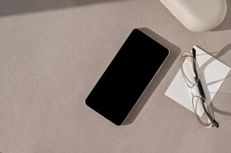 Minimal business brand template, flat lay. Mobile phone with empty black screen, eyeglasses, paper card on neutral beige background with natural sun light shadow.の写真素材