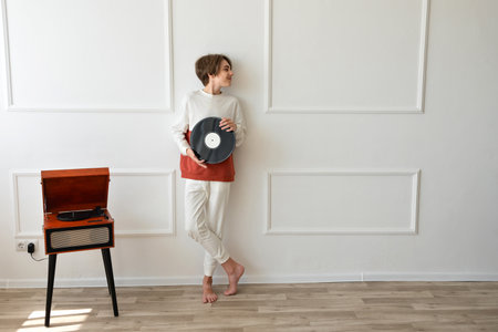 Cheerful handsome boy standing near turntable, smiling and holding vinyl record in hands, empty white wall background behind.の写真素材