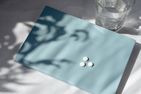Everyday health care morning routine aesthetic concept. Pills, vitamins or supplements on blue notebook, glass with water on white table with floral sunlight shadows.の写真素材