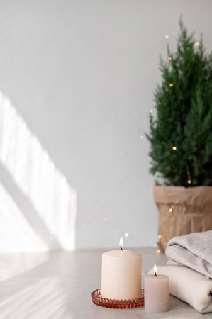 Scandinavian winter home interior decoration. Candles, juniper in pot, sweaters on beige table, empty white wall background with aesthetic sunlight shadows. Minimal white Christmas design template.の写真素材
