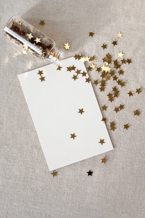 Blank paper card mockup with gold glitter star confetti scattered from bottle on neutral beige linen tablecloth background. Aesthetic New Year or birthday greeting postcard, party invitation template.の写真素材