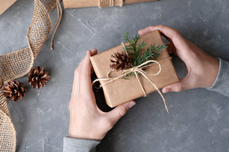 Person's hands holding brown crafted gift box decorated with pine branch and cone over gray table background. Sustainable aesthetic Christmas celebration, presents preparing concept.の写真素材