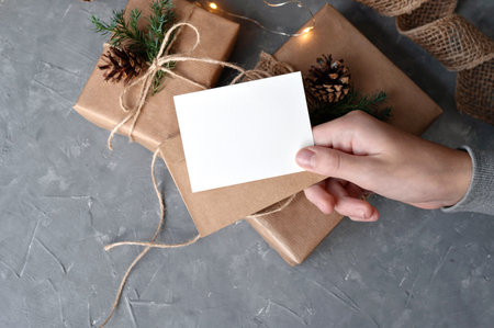 Christmas presents, gift boxes wrapped with brown crafted paper, pine cones and twig decor, on gray table background. Empty paper card mockup in hand. Gift tag, business card or invitation template.の写真素材