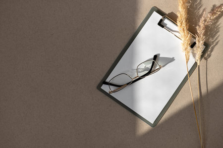 Blank business papers on clipboard and eyeglasses, dried meadow grass on neutral brown table with geometric sunlight shadows. Sustainable business, financial, corporate minimalist concept.の写真素材
