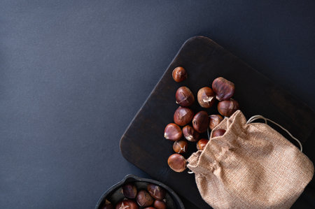 Edible roasted chestnuts in jute sack scattered on brown wooden board, on dark gray background with copy space. Top view, flat lay.の写真素材
