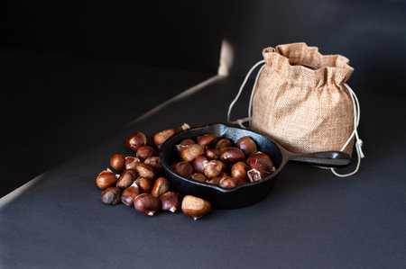 Pan with fried edible chestnuts, jute sack on black table background with sunlight ray. Traditional healthy organic autumn and winter food. Soft selective focus.の写真素材