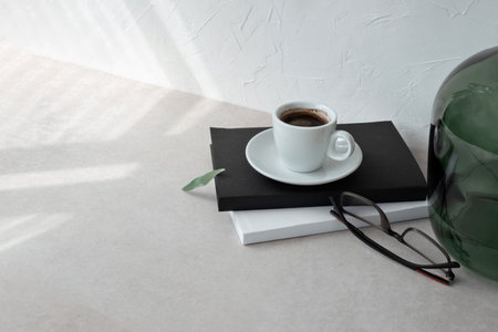 Cup with coffee drink, books, eyeglasses on beige table, empty white wall background with aesthetic sunlight pattern. Elegant business branding template.の写真素材