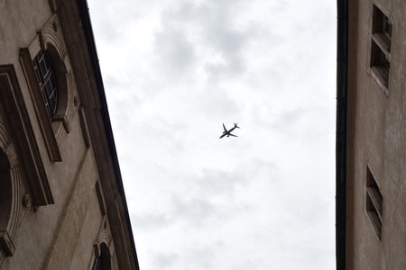 Small silhouette of Jet airplane flying high in cloudy sky above old town buildings. Travel and...