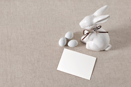 Minimalist Easter holiday greeting postcard, invitation or business card template, blank paper card mockup, ceramic rabbit figurine and small eggs on neutral taupe linen tablecloth background.の写真素材