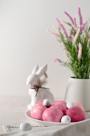 Traditional Easter food and table setting decor, pink dyed eggs on plate, rabbit figurine, spring flowers in vase on beige linen tablecloth, blurred background, soft natural light, selective focus.の写真素材