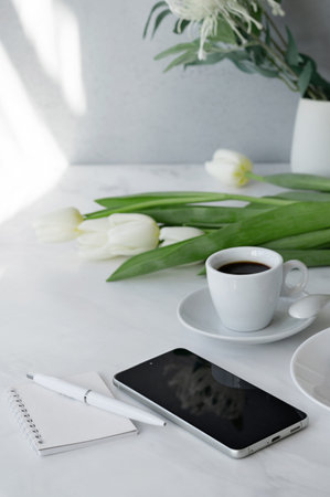 Elegant home office workspace, mobile phone, notebook, pen, cup of coffee and flowers on white table...