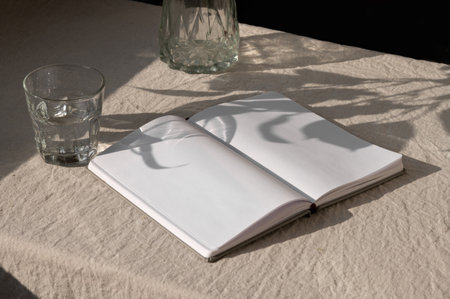 Tabletop with open book or notepad with blank sheets mockup, glass with water, vase on neutral beige...