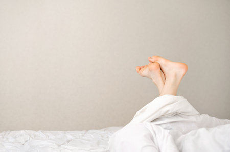 Person bare feet rise up in bed under white linen blanket, on empty neutral beige wall background, copy space, good sleep, comfortable sleeping bedding concept.の写真素材