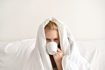 Morning coffee, hard morning wake up concept, young teenage girl covered by white blanket sitting on bed in neutral light bedroom, drinking coffee from white ceramic cup.の写真素材