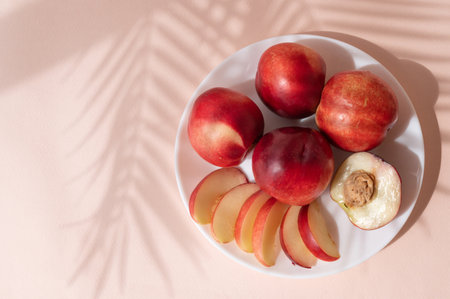 Whole and sliced sweet red nectarine fruits on plate on pink background with leaf shadows.の写真素材