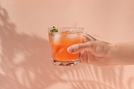 Hand holding glass with iced summer refreshing drink with peach fruits and ice, on pastel peachy...