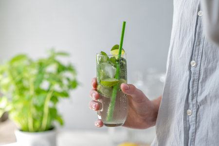 Person hand holding glass with mojito cocktail drink with ice, mint, lime and green drinking straw...