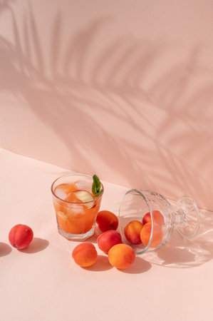 Glass with iced refreshing peach juice drink, peaches and apricots scattered on bright peachy pink background with abstract floral sun light shadows.の写真素材