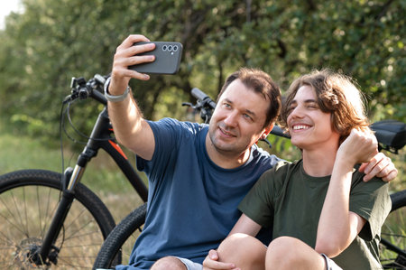 Single father and teenage son taking photo on mobile phone, while have a rest after cycling, sitting in nature with bicycles, talking and smiling, family active lifestyle.の写真素材