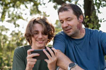 Teenager boy and his father sitting in nature and looking at smartphone screen, talking and smiling.の写真素材