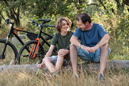 Father and teenager son resting together outdoor in nature, sitting near bicycles, talking and smiling.の写真素材
