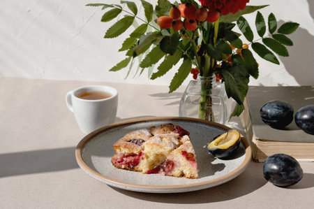 Plate with piece of plum pie, fresh plums, cup with tea, vase with autumn flowers bouquet on beige table background, seasonal fall sweet food for Thanksgiving holiday dinner.の写真素材