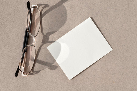 Blank white paper card mockup, glasses on neutral taupe brown desk background with aesthetic natural sunlight shadows, minimalist business card design template.の写真素材