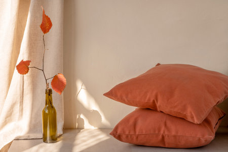 Cozy autumn home interior decor, warm terracotta red pillows and vase with fall orange leaves, linen beige curtain with natural sunlight, empty neutral wall background.の写真素材