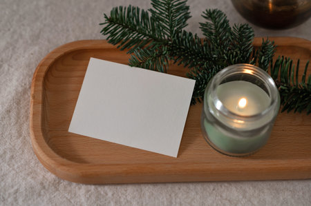 Blank paper card mockup, green candle with soft dim light, fir branch on wooden tray on table, cozy aesthetic winter home still life, template for postcard or invitation.の写真素材