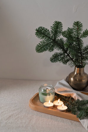 Aesthetic Christmas still life in Scandinavian style, cozy winter home decor, fir tree branch in vase, candles with dim light on wooden tray on table with linen tablecloth, empty gray wall background.の写真素材