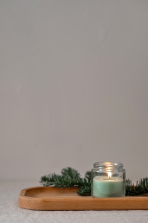 Scented green candle with dim light and fir branch on wooden tray on table, empty neutral gray beige wall background, aesthetic minimalist natural home still life, empty copy space.の写真素材