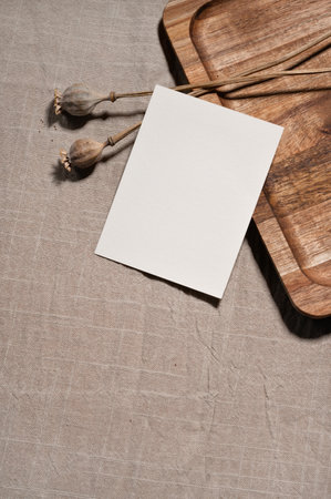 Empty paper card mockup, dried poppy flowers on brown natural wooden tray and neutral beige linen tablecloth background, elegant wedding invitation or greeting postcard design template.の写真素材