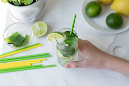 Fresh summer drink on home kitchen table with green and yellow lime, mint and drinking straws, hand holding glass with iced mojito cocktail beverage, lifestyle.の写真素材