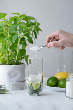 Pouring sugar in glass with lime and mint, kitchen table with mojito ingredients, fresh summer drink making process.の写真素材