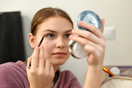 Teenage girl eye makeup, young woman looking at small compact mirror and do eye makeup with eyeliner and eyeshadow, authentic lifestyleの写真素材