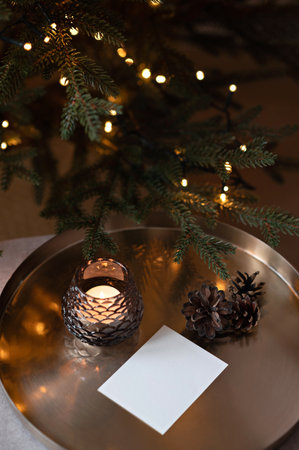 Blank paper card mockup, candle light, pine cone on cooper tray, blurred Christmas tree lights bokeh on background, aesthetic holiday postcard or invitation design template in dark brown green tonesの写真素材