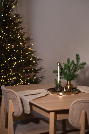 Aesthetic Christmas holiday home interior, beige wooden table with chairs, candle light and vase on cooper tray, linen table runner, Christmas tree shining light bokeh on background, evening lightの写真素材
