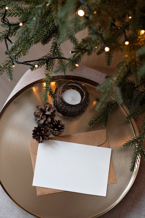 Blank paper card mockup, envelope, candle and pine cone on golden tray near blurred Christmas tree with shining lights, aesthetic dark moody background for holiday business brandingの写真素材
