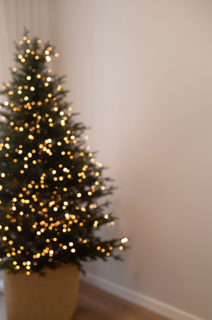 Blurred defocused Christmas tree with shining lights bokeh near empty neutral beige wall background, lifestyle real home interior, dark mood simple holiday backgroundの写真素材